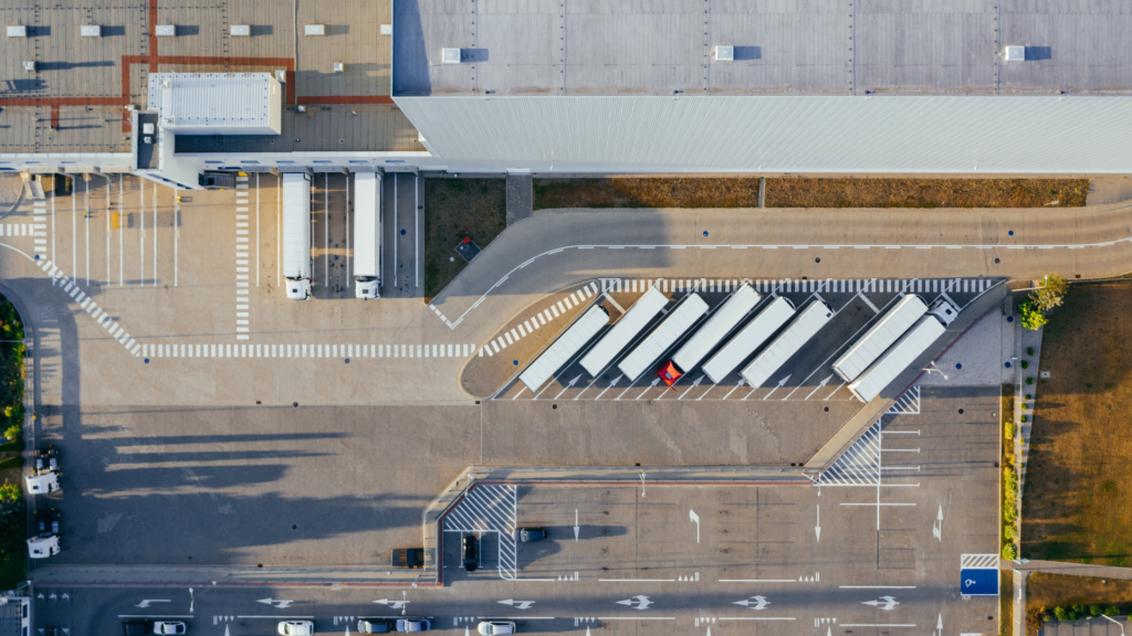 Aerial view of a logistics warehouse with semi-trailers parked at loading docks and in a diagonal truck parking area, with a car park and access road below.